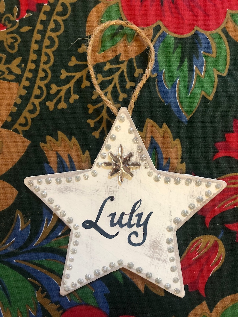 May include: White star-shaped Christmas ornament with a silver snowflake and the name "Luly" written in black calligraphy.