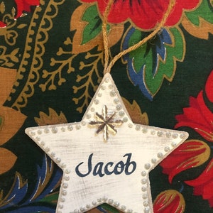 May include: White wooden star ornament with a silver snowflake and the name Jacob written in black paint.