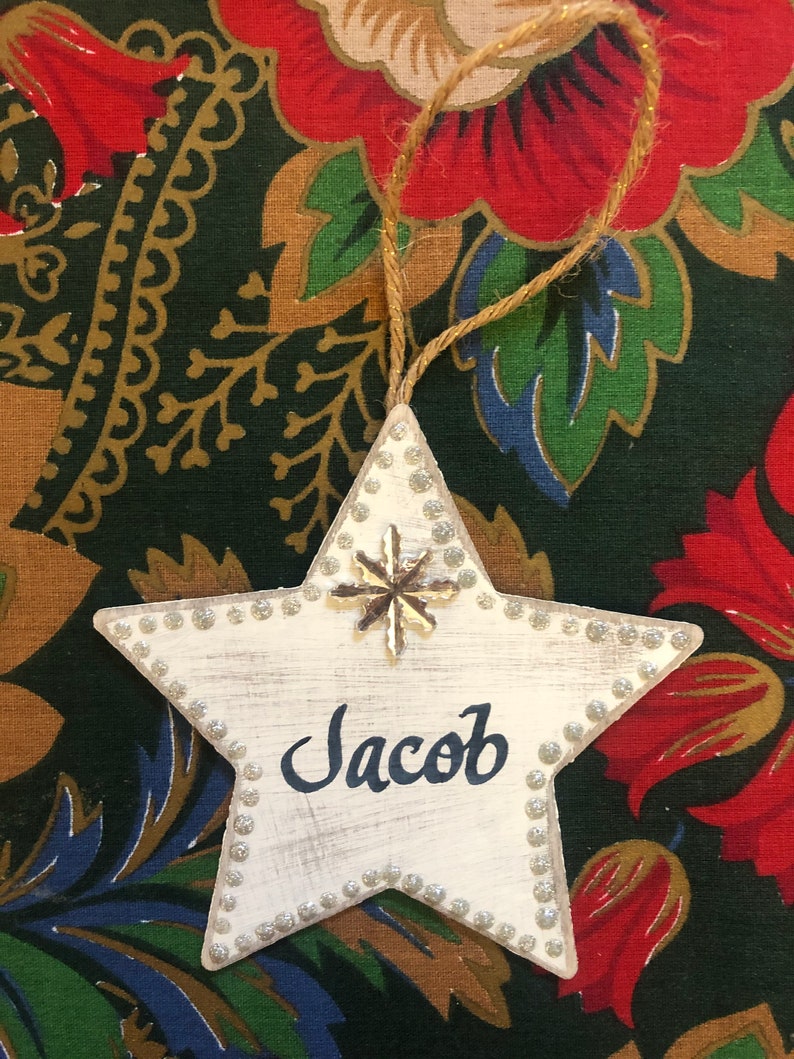 May include: White wooden star ornament with a silver snowflake and the name Jacob written in black paint.