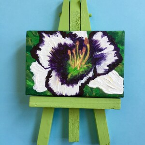 Puede incluir: Una peque&ntilde;a pintura de una flor blanca con un centro morado en un caballete verde.