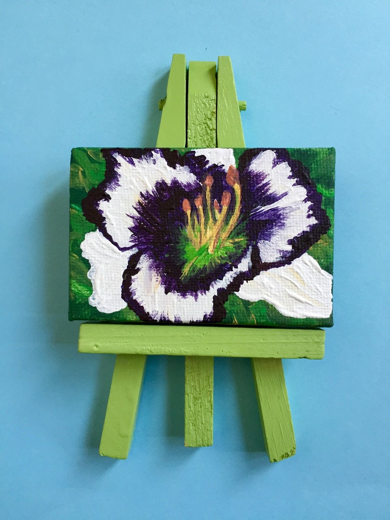 Puede incluir: Una peque&ntilde;a pintura de una flor blanca con un centro morado en un caballete verde.
