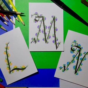 May include: Three cards featuring hand-lettered calligraphy of the letters L, M, and N. Each letter is adorned with a vine of flowers. The L is decorated with yellow flowers, the M with purple flowers, and the N with blue flowers.