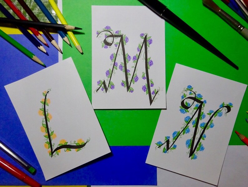 May include: Three cards featuring hand-lettered calligraphy of the letters L, M, and N. Each letter is adorned with a vine of flowers. The L is decorated with yellow flowers, the M with purple flowers, and the N with blue flowers.