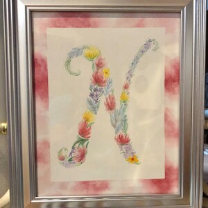 May include: A framed watercolor painting of the letter "N" made of flowers. The flowers are in shades of pink, yellow, blue, and green. The background is a watercolor wash of pink and white.