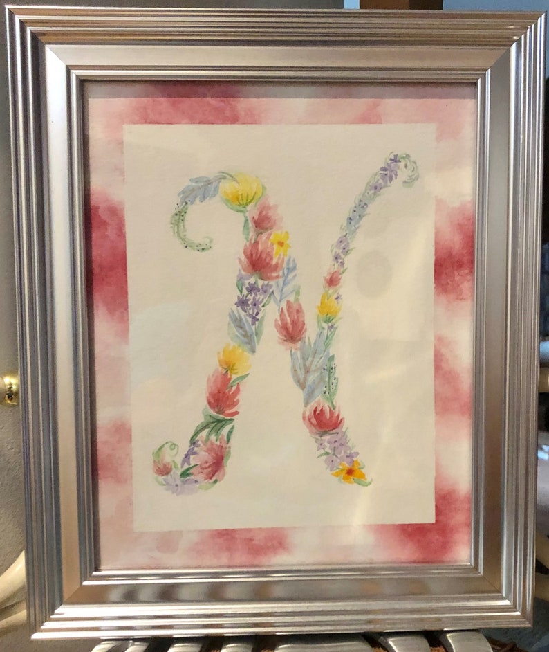 May include: A framed watercolor painting of the letter "N" made of flowers. The flowers are in shades of pink, yellow, blue, and green. The background is a watercolor wash of pink and white.