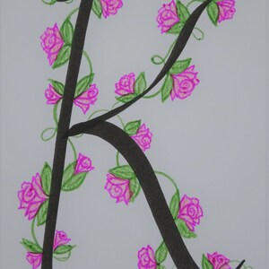 May include: A black calligraphic letter K with a vine of pink roses winding around it. The roses have green leaves and stems.