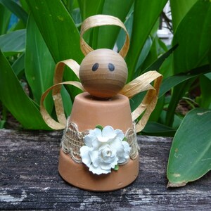 K&ouml;nnte beinhalten: Eine kleine Engelfigur aus einem Terrakotta-Topf mit einem Holzkopf, braunen Haaren und einer wei&szlig;en Blume. Der Engel tr&auml;gt ein braunes Leinenband und Spitze.