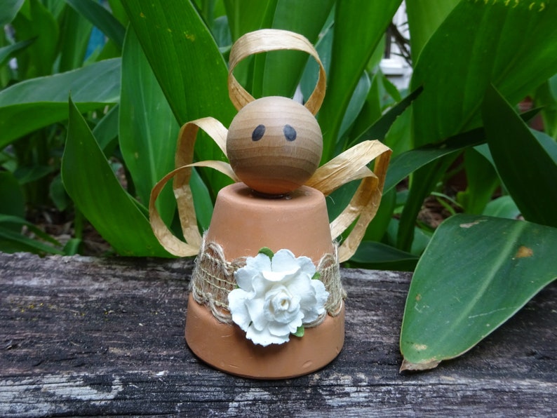 K&ouml;nnte beinhalten: Eine kleine Engelfigur aus einem Terrakotta-Topf mit einem Holzkopf, braunen Haaren und einer wei&szlig;en Blume. Der Engel tr&auml;gt ein braunes Leinenband und Spitze.
