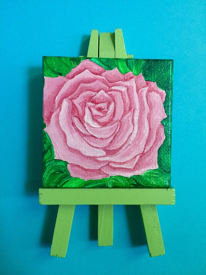 Puede incluir: Una rosa rosa pintada en un lienzo peque&ntilde;o, exhibida en un caballete verde.