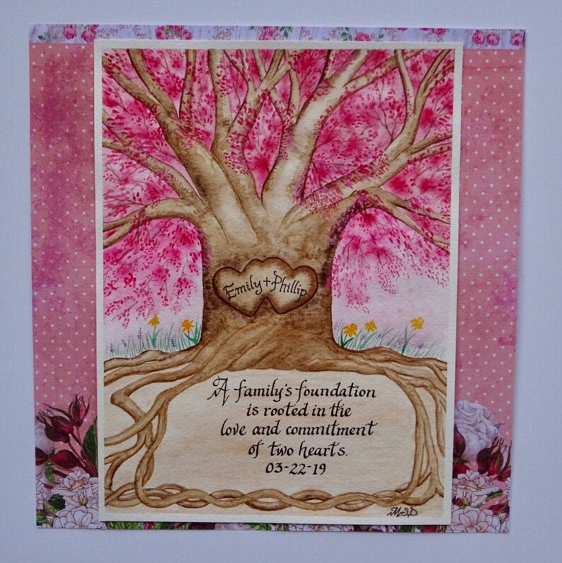May include: A watercolor painting of a tree with pink blossoms. Two intertwined hearts are carved into the trunk with the names "Emily + Phillip" inscribed. The roots of the tree are visible and the text "A family's foundation is rooted in the love and commitment of two hearts. 03-22-19" is written below the tree.