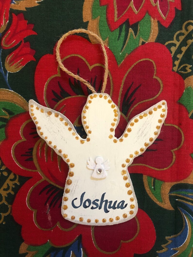 May include: White wooden angel ornament with gold glitter accents and the name "Joshua" written in black paint. The ornament has a small white angel with a rhinestone in the center.