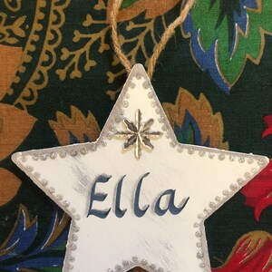 May include: White star-shaped Christmas ornament with a silver glitter border and a silver snowflake charm. The ornament is personalized with the name "Ella" in blue paint.