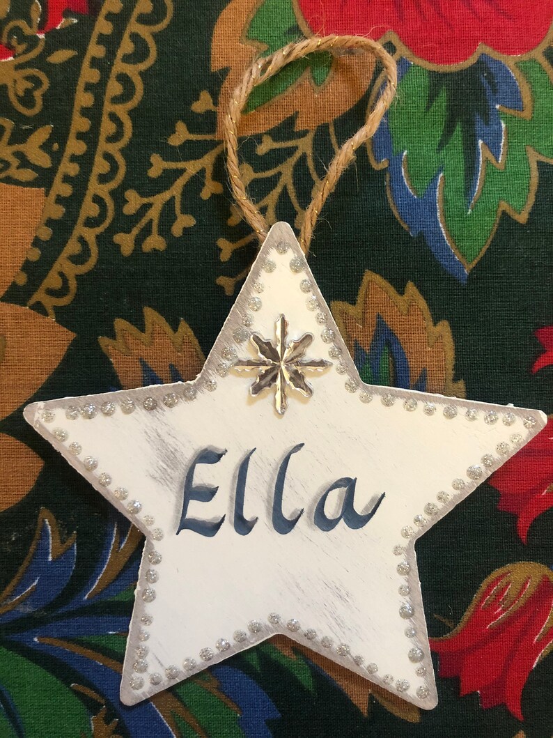 May include: White star-shaped Christmas ornament with a silver glitter border and a silver snowflake charm. The ornament is personalized with the name "Ella" in blue paint.