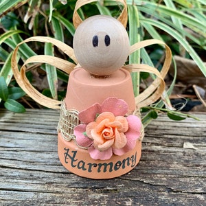 K&ouml;nnte beinhalten: Eine kleine Engelfigur aus einem Terrakotta-Topf mit einer rosa Blume und dem Wort "Harmonie" auf der Vorderseite. Der Engel hat einen Holzkopf mit schwarzen Augen und stroh&auml;hnliche Fl&uuml;gel.