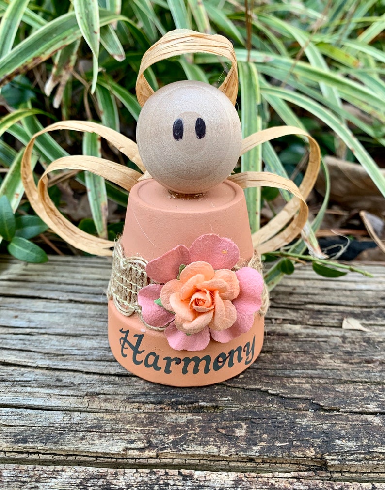 K&ouml;nnte beinhalten: Eine kleine Engelfigur aus einem Terrakotta-Topf mit einer rosa Blume und dem Wort "Harmonie" auf der Vorderseite. Der Engel hat einen Holzkopf mit schwarzen Augen und stroh&auml;hnliche Fl&uuml;gel.