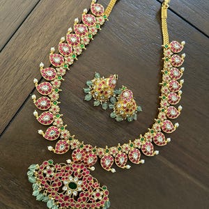May include: A gold-toned jewelry set featuring a necklace, earrings, and a pendant. The necklace and pendant have a paisley design with red and white accents, green details, and small white pearls. The earrings have a similar design with green beads.