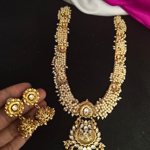 May include: Gold-toned necklace and earrings set. The necklace features a pendant with clear stones and a floral design, and is adorned with small, white pearls. The earrings are bell-shaped with a floral design.