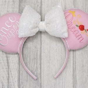 May include: Pink Minnie Mouse ears with a white bow and embroidered text that says "Once Upon a Dream" with a crown and rose design.