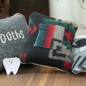 Puede incluir: Tres pequeños cojines de tela con estampado gris y negro. Un cojín tiene la palabra "Tooths" bordada. Un diente de papel blanco con una cara sonriente está delante de los cojines.