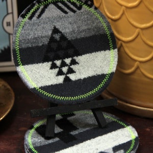 Puede incluir: Un juego de cuatro posavasos redondos con un patrón geométrico en negro, blanco y gris. Los posavasos están hechos de un material suave y peludo y tienen un borde cosido verde.