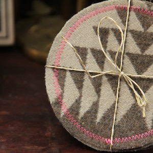 Puede incluir: Un juego de cuatro posavasos marrones y blancos con un patrón geométrico. Los posavasos están atados con una cuerda blanca y tienen un borde cosido rosa.