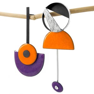 Puede incluir: Un par de pendientes geométricos con detalles en naranja, morado, negro y plata. Los pendientes están hechos de metal y tienen un diseño moderno.