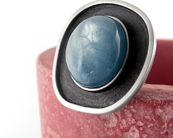May include: A ring featuring a smooth, round, light blue cabochon stone set in a dark, textured, circular metal frame with a polished silver-toned outer edge. The design highlights a contrast between the blue stone and the dark metal.