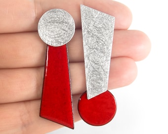 Peut inclure: Une paire de boucles d'oreilles dépareillées aux formes géométriques. L'une possède un haut circulaire argenté avec une base trapézoïdale rouge. L'autre a un haut trapézoïdal argenté avec une base circulaire rouge. Les parties argentées sont brossées.