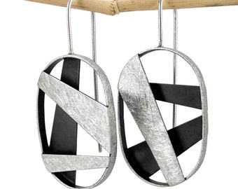 May include: A pair of oval-shaped drop earrings featuring a silver-toned frame with a textured, brushed finish. The interior of each earring contains intersecting black metal strips that create a geometric, open-work design with long hooks.