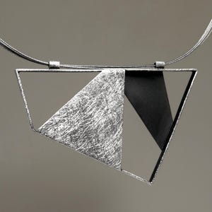 May include: A geometric pendant necklace featuring an open trapezoidal frame. The interior contains a textured silver triangular plate with a brushed finish alongside a smooth, solid black triangular plate, suspended from a thin silver wire cord.