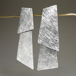 Puede incluir: Pendientes geométricos plateados con un acabado de metal cepillado texturizado. Cada pendiente presenta dos piezas trapezoidales superpuestas. Los pendientes cuelgan de un fino alambre dorado, creando un diseño moderno y minimalista.