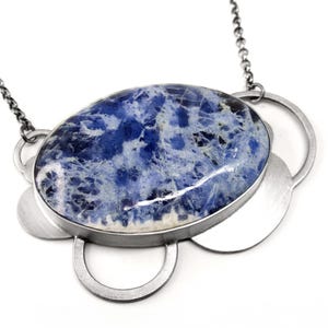 Puede incluir: Un collar con una gran piedra de sodalita ovalada de color azul y blanco engastada en un marco de metal cepillado. El colgante está decorado con varios discos metálicos circulares superpuestos y cuelga de una fina cadena de bolas oscura.