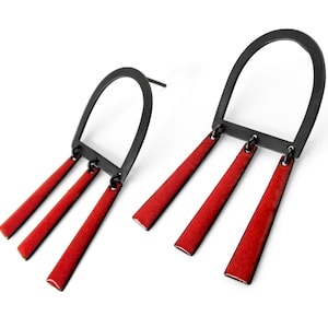 Puede incluir: Un par de pendientes de metal negro con detalles de esmalte rojo. Cada pendiente tiene una forma de arco curvo con tres tiras de esmalte rojo colgantes.