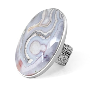 Puede incluir: Un anillo con un gran cabochon ovalado de ágata bandeada en tonos blancos, grises y rosa suave. La piedra está engastada en un bisel liso de tono plateado, y la banda del anillo tiene un acabado metálico distintivo, rugoso y texturizado.