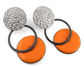 May include: A pair of earrings featuring a textured silver-tone circular top piece connected to a thin black metal ring, which holds a smooth, vibrant orange circular disc at the bottom. The design combines contrasting metallic finishes and colors.