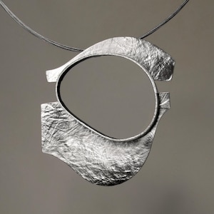 Puede incluir: Un collar con colgante plateado con un diseño abstracto texturizado. El colgante presenta una forma ovalada abierta con una superficie texturizada y está suspendido de un fino cordón oscuro. El diseño es moderno y minimalista.