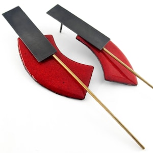 Puede incluir: Un par de pendientes que presentan un segmento curvo de esmalte rojo brillante unido a una barra rectangular horizontal de color negro mate. Una varilla de metal larga y delgada de tono dorado se extiende diagonalmente.