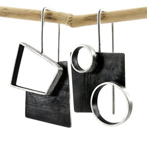 Pode incluir: Um par de brincos assimétricos com ganchos de metal prateado. Um brinco apresenta uma moldura trapezoidal prateada sobre uma placa retangular escura. O outro brinco tem uma placa quadrada escura com dois anéis circulares prateados.