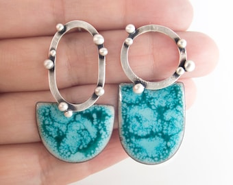 May include: A pair of mismatched earrings featuring oxidized silver frames with small spherical accents. One earring has an oval frame, and the other has a circular frame. Both are attached to semi-circular dangles with a mottled teal enamel finish.