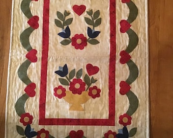May include: A rectangular, cream-colored quilted wall hanging with a floral design. The central panel features a red-bordered rectangle with floral and heart motifs. A green, wavy border with red hearts and flowers frames the central design.