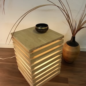 Könnte beinhalten: Ein Holz-Beistelltisch mit einer hellen Holzoptik und einem integrierten Licht. Der Tisch hat eine quadratische Tischplatte und besteht aus übereinander gestapelten Holzlatten. Eine kleine schwarze Klangschale steht auf der Tischplatte.