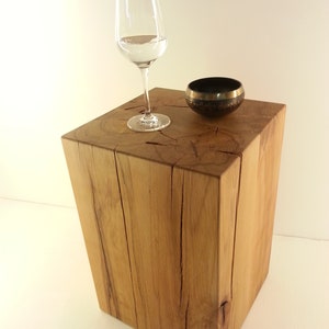 Puede incluir: Una mesa auxiliar de madera marrón claro con una copa de vino transparente llena de agua y un pequeño cuenco negro y dorado encima. La mesa tiene vetas de madera natural y forma cuadrada.