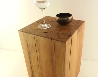 Puede incluir: Una mesa auxiliar de madera marrón claro con una copa de vino transparente llena de agua y un pequeño cuenco negro y dorado encima. La mesa tiene vetas de madera natural y forma cuadrada.