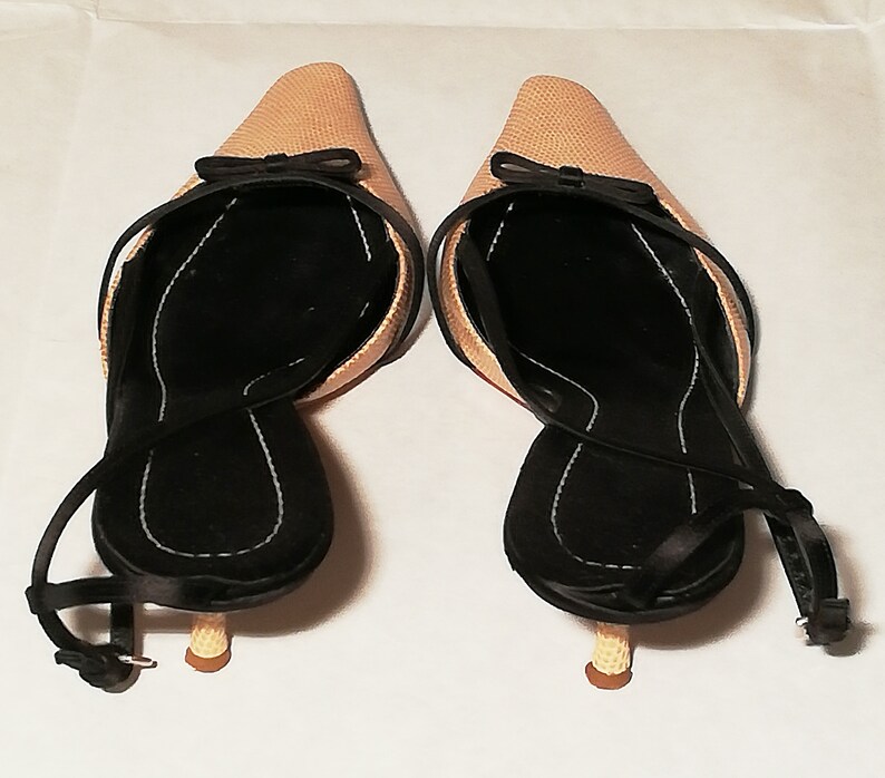 Puede incluir: Un par de zapatos de tac&oacute;n beige con tiras traseras, puntera afilada y peque&ntilde;os lazos negros. Los zapatos tienen plantillas negras y correas en los tobillos. Los tacones son de color marr&oacute;n claro.