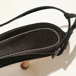 Puede incluir: Primer plano de una sandalia de ante negro con una fina correa en el tobillo. La sandalia presenta un tac&oacute;n texturizado en forma de cono y un borde cosido en blanco. La correa se asegura con una peque&ntilde;a hebilla plateada. El dise&ntilde;o general es elegante y minimalista.