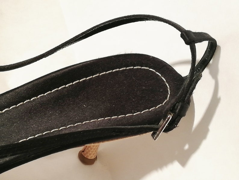 Puede incluir: Primer plano de una sandalia de ante negro con una fina correa en el tobillo. La sandalia presenta un tac&oacute;n texturizado en forma de cono y un borde cosido en blanco. La correa se asegura con una peque&ntilde;a hebilla plateada. El dise&ntilde;o general es elegante y minimalista.