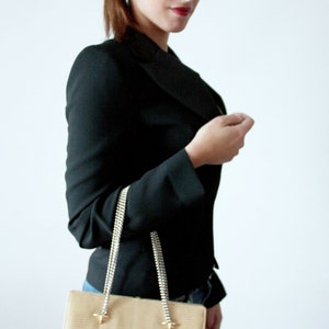 Puede incluir: Una mujer con un blazer negro y gafas de sol sostiene un bolso de mano beige con una correa de cadena plateada.