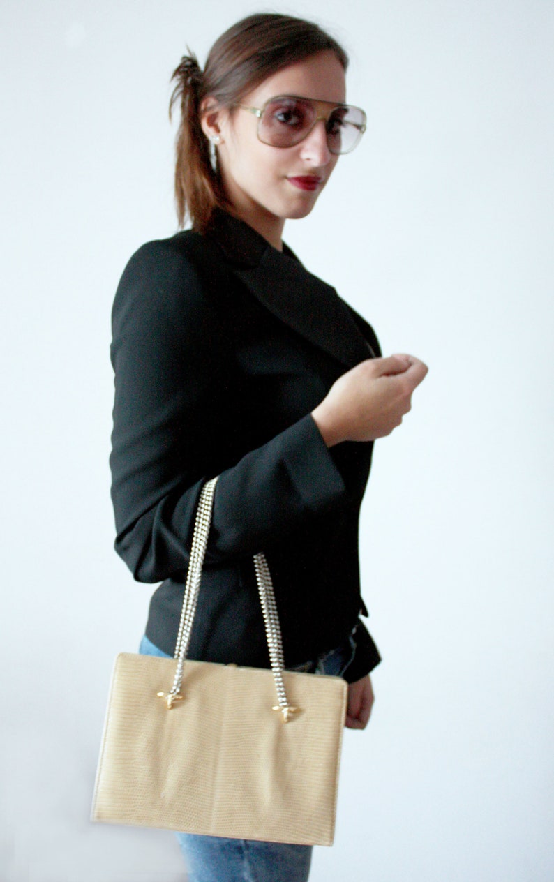 Puede incluir: Una mujer con un blazer negro y gafas de sol sostiene un bolso de mano beige con una correa de cadena plateada.