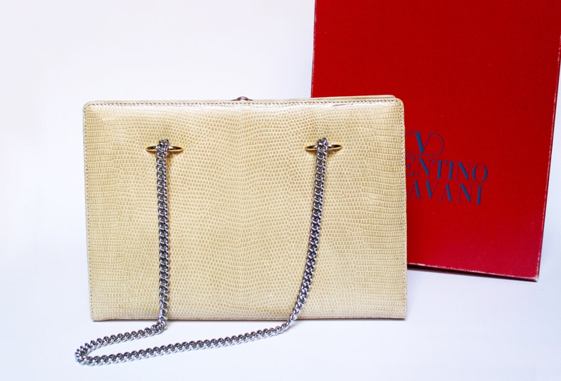 Puede incluir: Un bolso de mano de cuero color crema con una correa de cadena plateada y herrajes dorados. El bolso tiene un patr&oacute;n texturizado, similar a la piel de serpiente. El bolso est&aacute; sobre una superficie blanca. El logotipo de Valentino Garavani es visible en la caja roja detr&aacute;s del bolso.