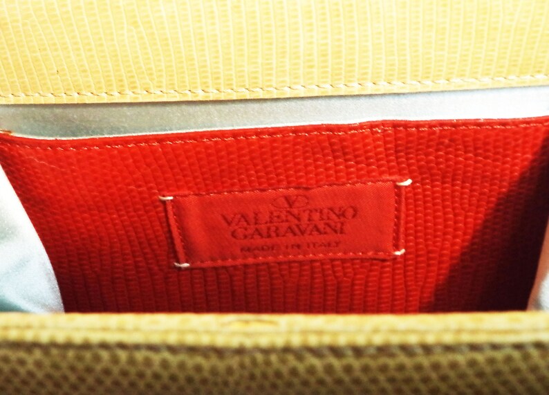 Puede incluir: Primer plano del interior de un bolso de mano de cuero rojo con una etiqueta de Valentino Garavani. La etiqueta es blanca con texto negro y un peque&ntilde;o logotipo de coraz&oacute;n rojo.